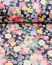 Japanese chiyogami paper featuring pink, yellow, purple, and white cherry blossoms and purple yukiwas on a black background with metallic gold accents.