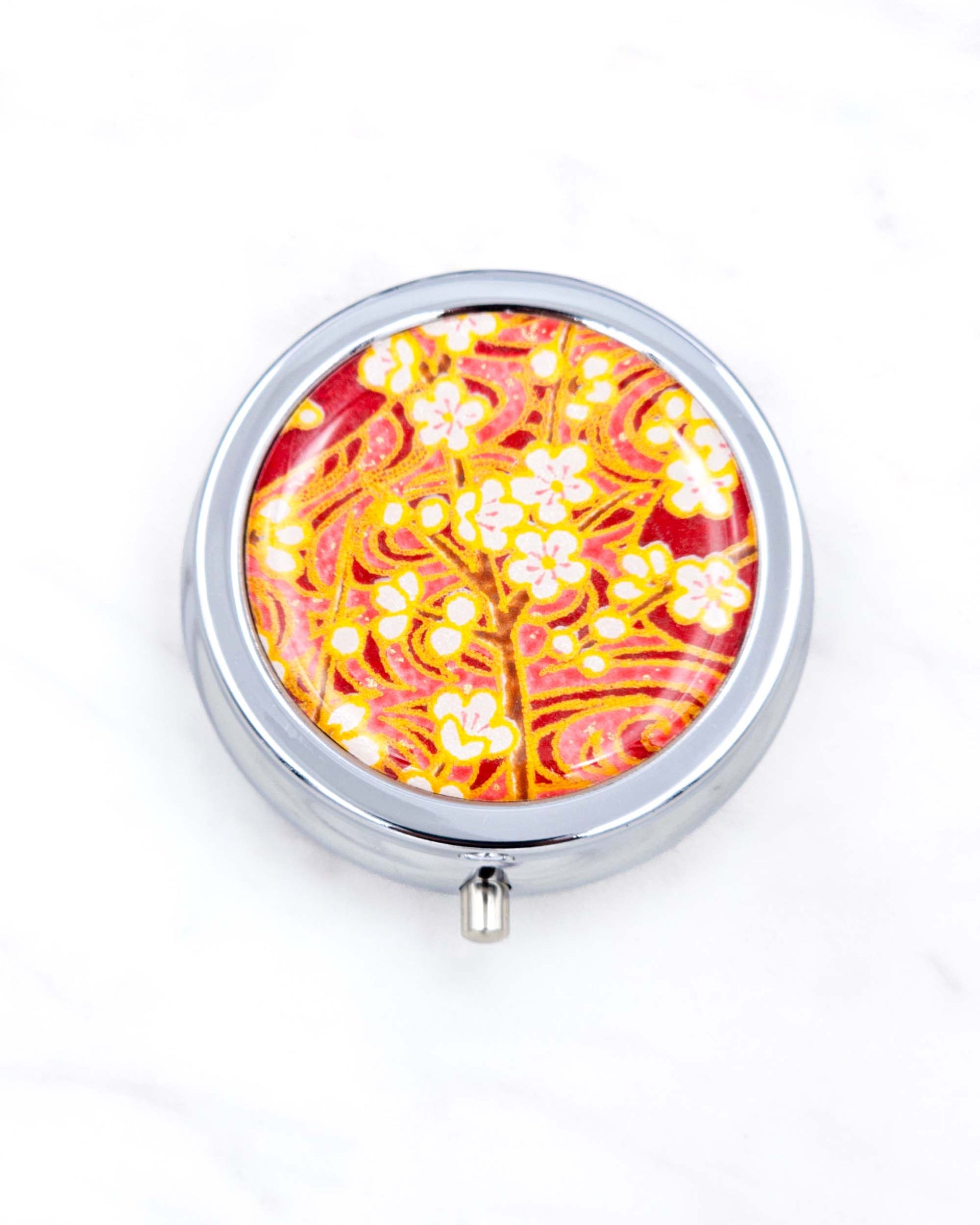 Silver metal pill box featuring white plum blossom branches on a maroon red background with metallic gold accents.