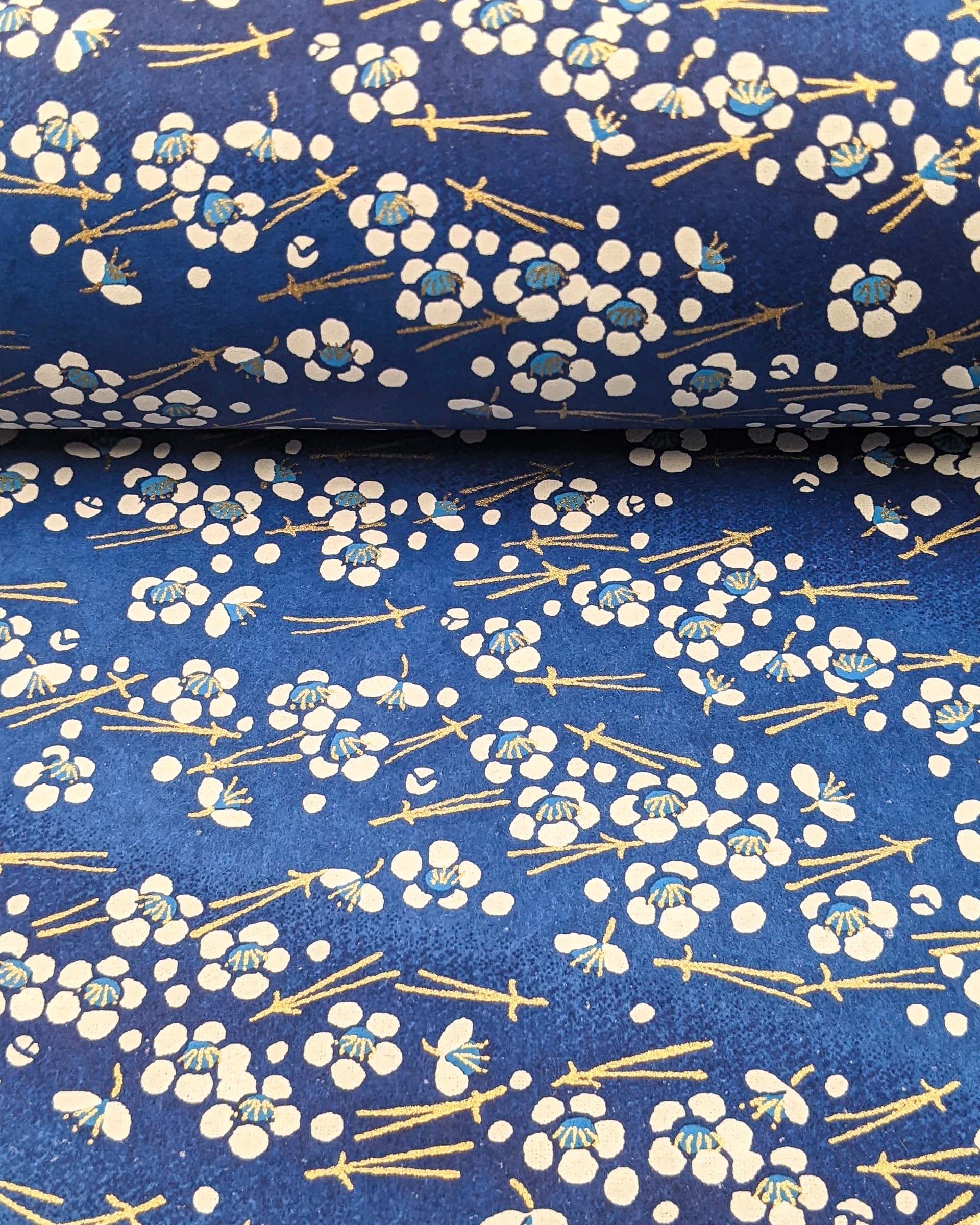 Japanese chiyogami featuring small white plum blossoms & matsuba (pine needles) on a blue background with metallic gold accents.