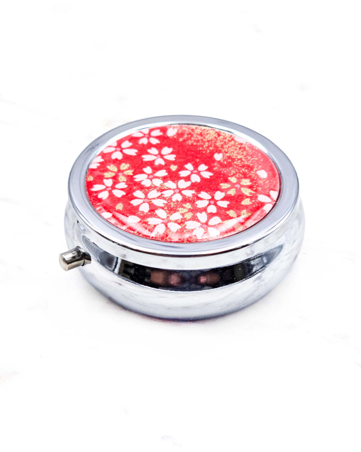 Round silver metal pill box featuring small white and gold cherry blossoms on a red background with metallic gold details.
