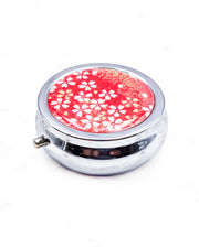 Round silver metal pill box featuring small white and gold cherry blossoms on a red background with metallic gold details.