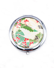 Round silver metal compact mirror featuring white flying cranes and red and white cherry blossoms on a green background with metallic gold details.