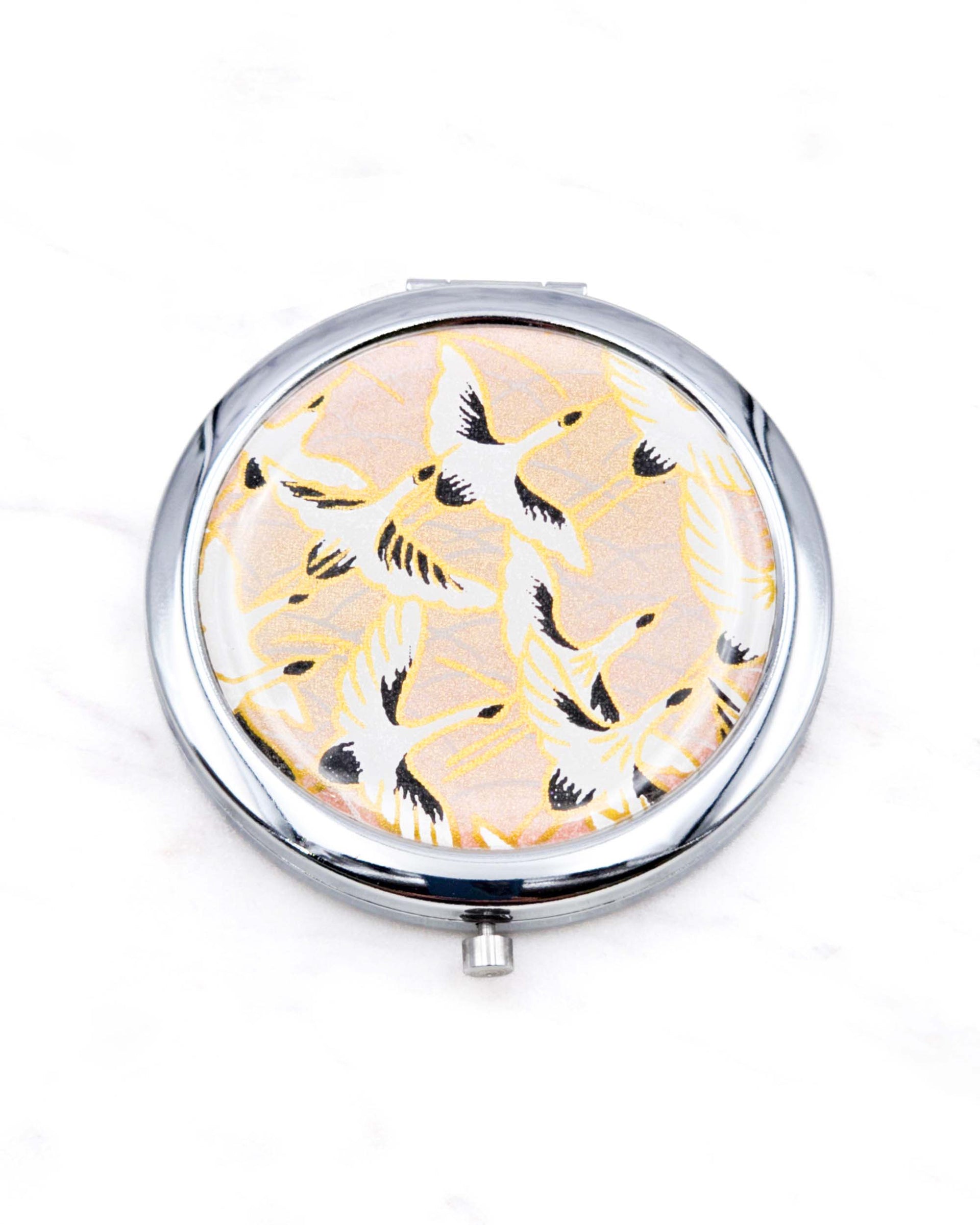 Round silver metal compact mirror featuring white flying cranes on a light pink background with metallic gold accents.