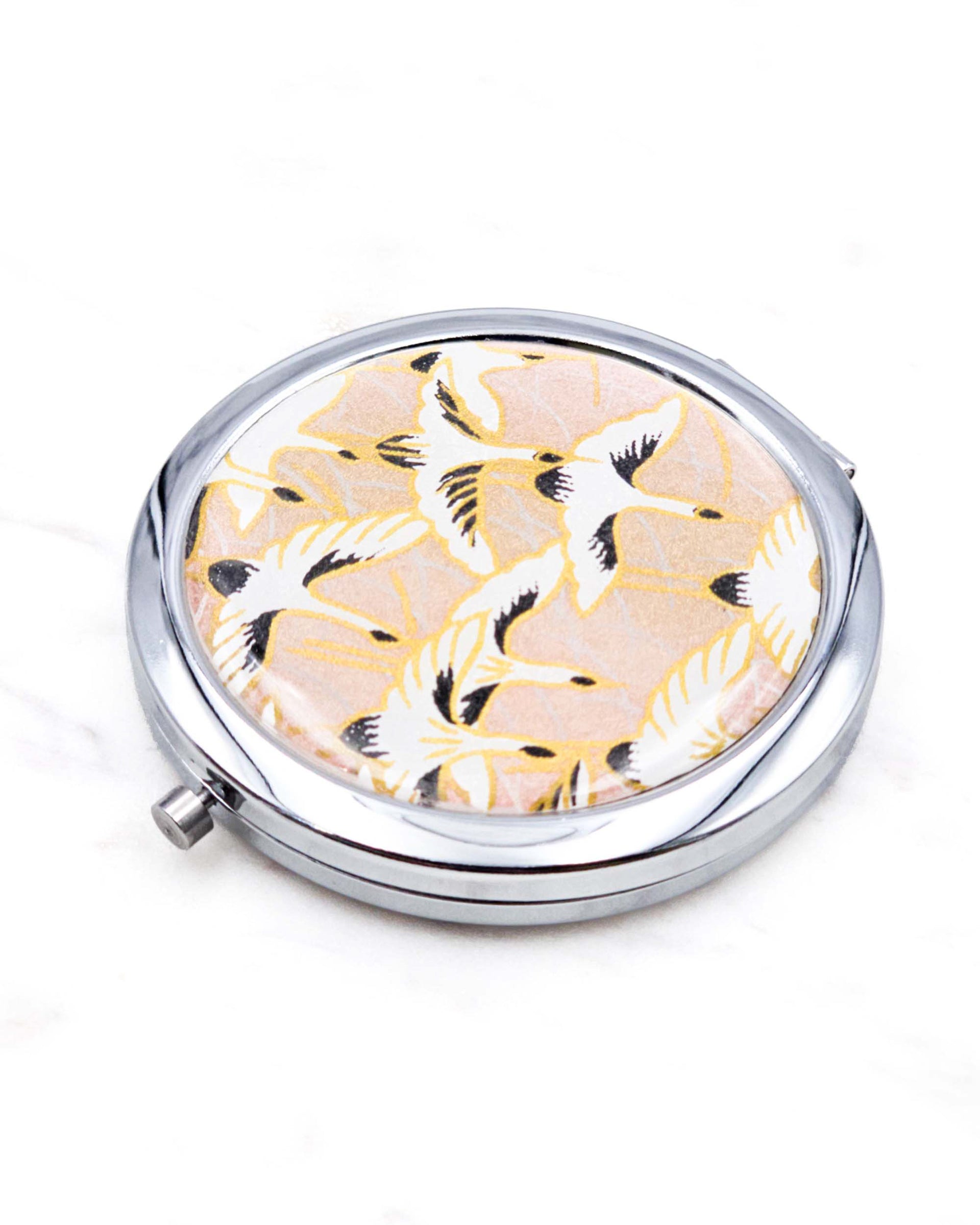 Round silver metal compact mirror featuring white flying cranes on a light pink background with metallic gold accents.