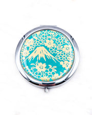 Round silver metal compact mirror featuring metallic gold Mt Fuji and cherry blossoms on a teal blue background.