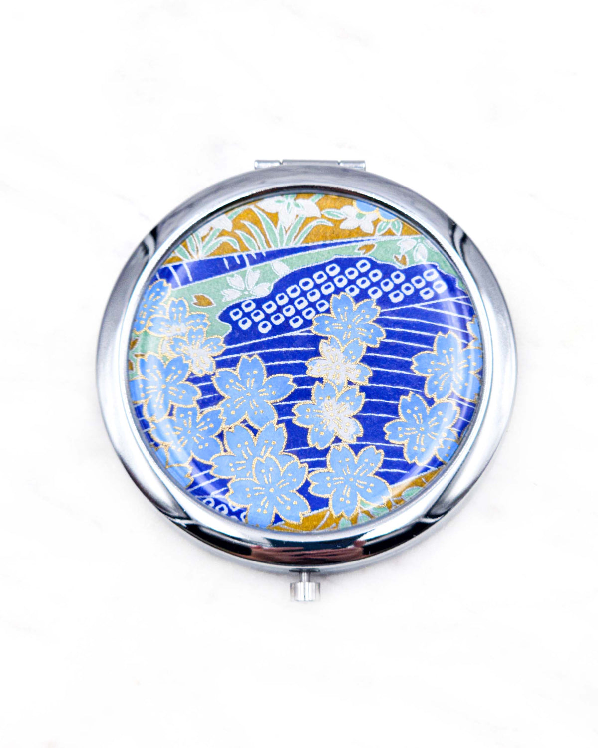 Round silver metal compact mirror featuring light blue cherry blossoms with metallic gold details on a dark blue background with flowing white lines and traditional Japanese patterns.