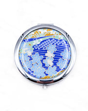 Round silver metal compact mirror featuring light blue cherry blossoms with metallic gold details on a dark blue background with flowing white lines and traditional Japanese patterns.