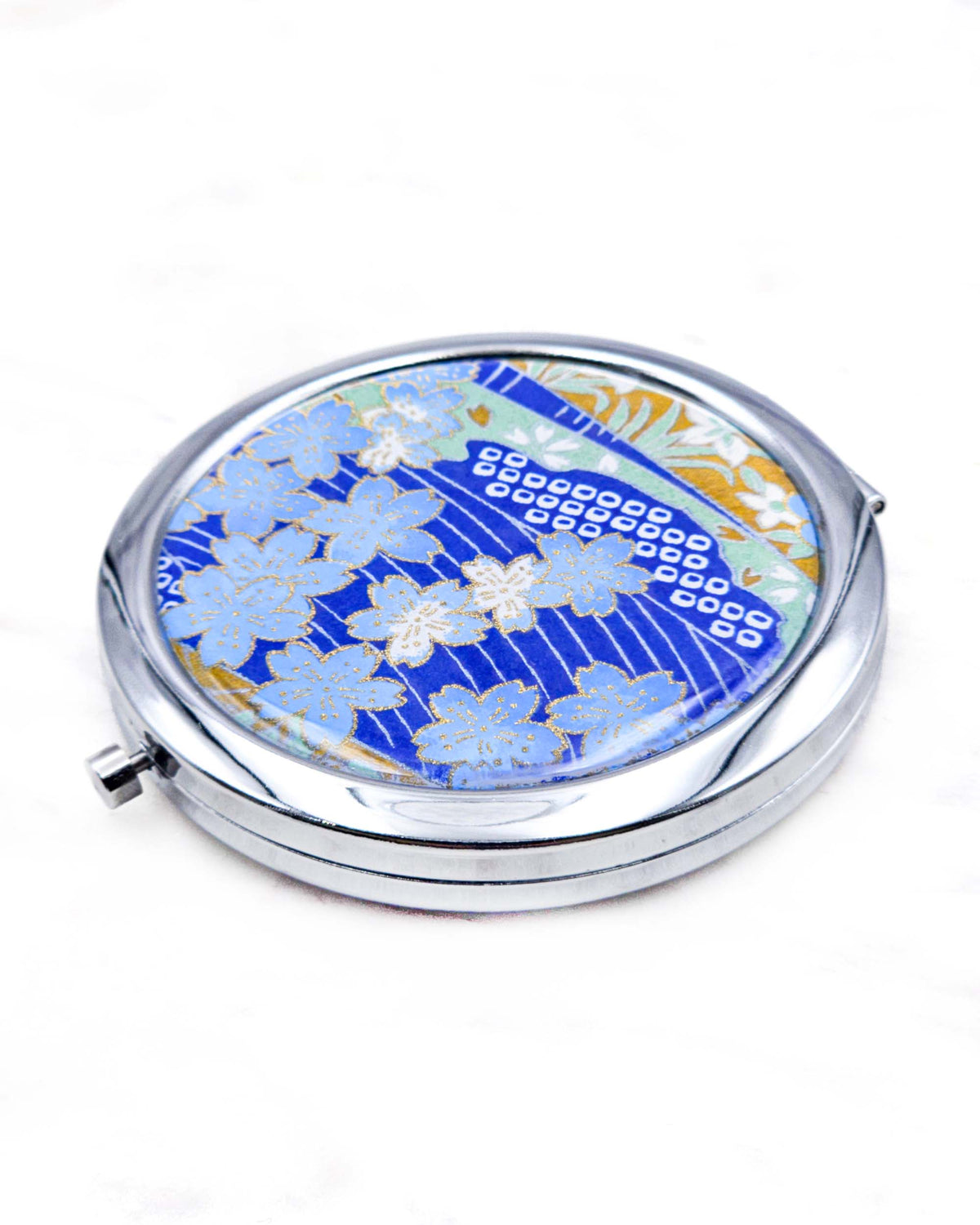 Round silver metal compact mirror featuring light blue cherry blossoms with metallic gold details on a dark blue background with flowing white lines and traditional Japanese patterns.
