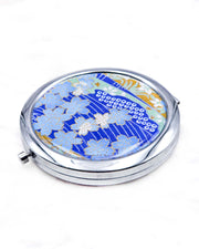 Round silver metal compact mirror featuring light blue cherry blossoms with metallic gold details on a dark blue background with flowing white lines and traditional Japanese patterns.