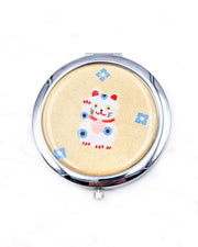 Round silver metal compact mirror featuring a white, red, and blue maneki-neko (lucky cat) with blue hanabishi (flower diamond) motifs on a metallic gold background.