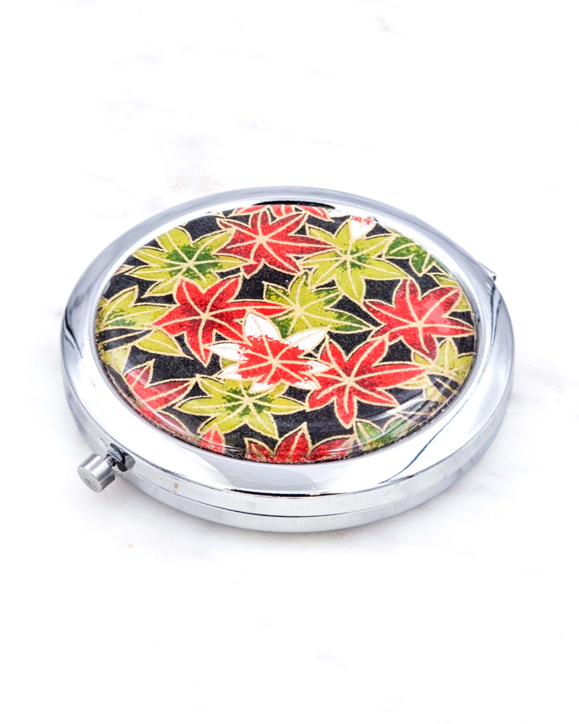 Silver metal compact mirror featuring green, red, and white maples leaves on a black background with metallic gold accents.