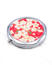 Round silver metal compact mirror featuring pink and white cherry blossoms on a red asanoha (hemp leaf) patterned background with metallic gold details.