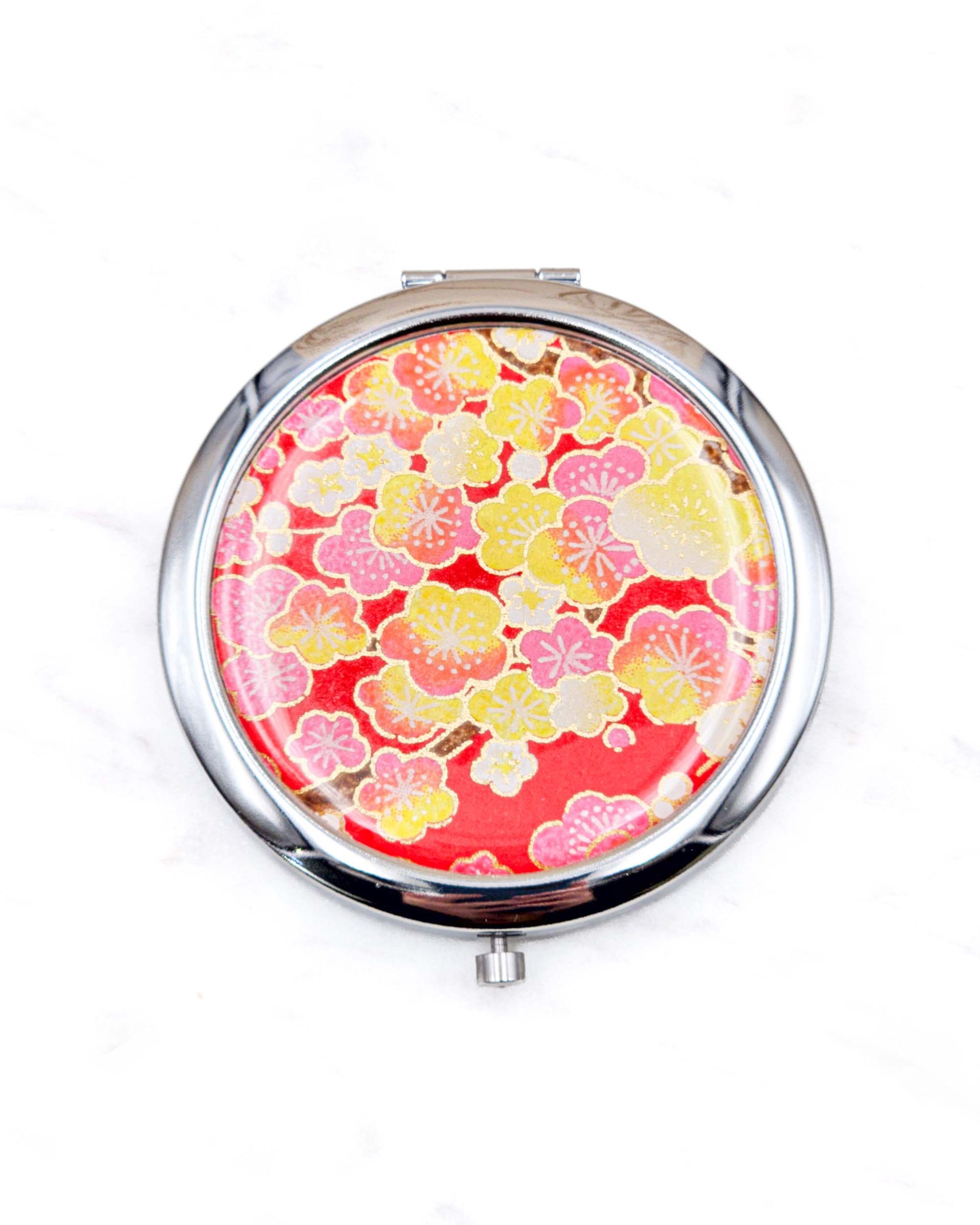 Round silver metal compact mirror featuring pink, yellow, and white plum blossoms on a red background with metallic gold details.