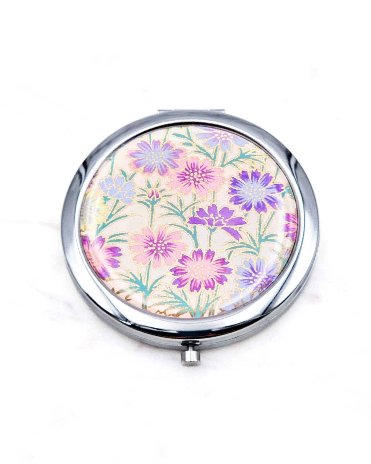 Round silver metal compact mirror featuring purple flower stalks on a light pink background with metallic gold accents.