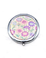 Round silver metal compact mirror featuring purple flower stalks on a light pink background with metallic gold accents.