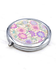 Round silver metal compact mirror featuring purple flower stalks on a light pink background with metallic gold accents.