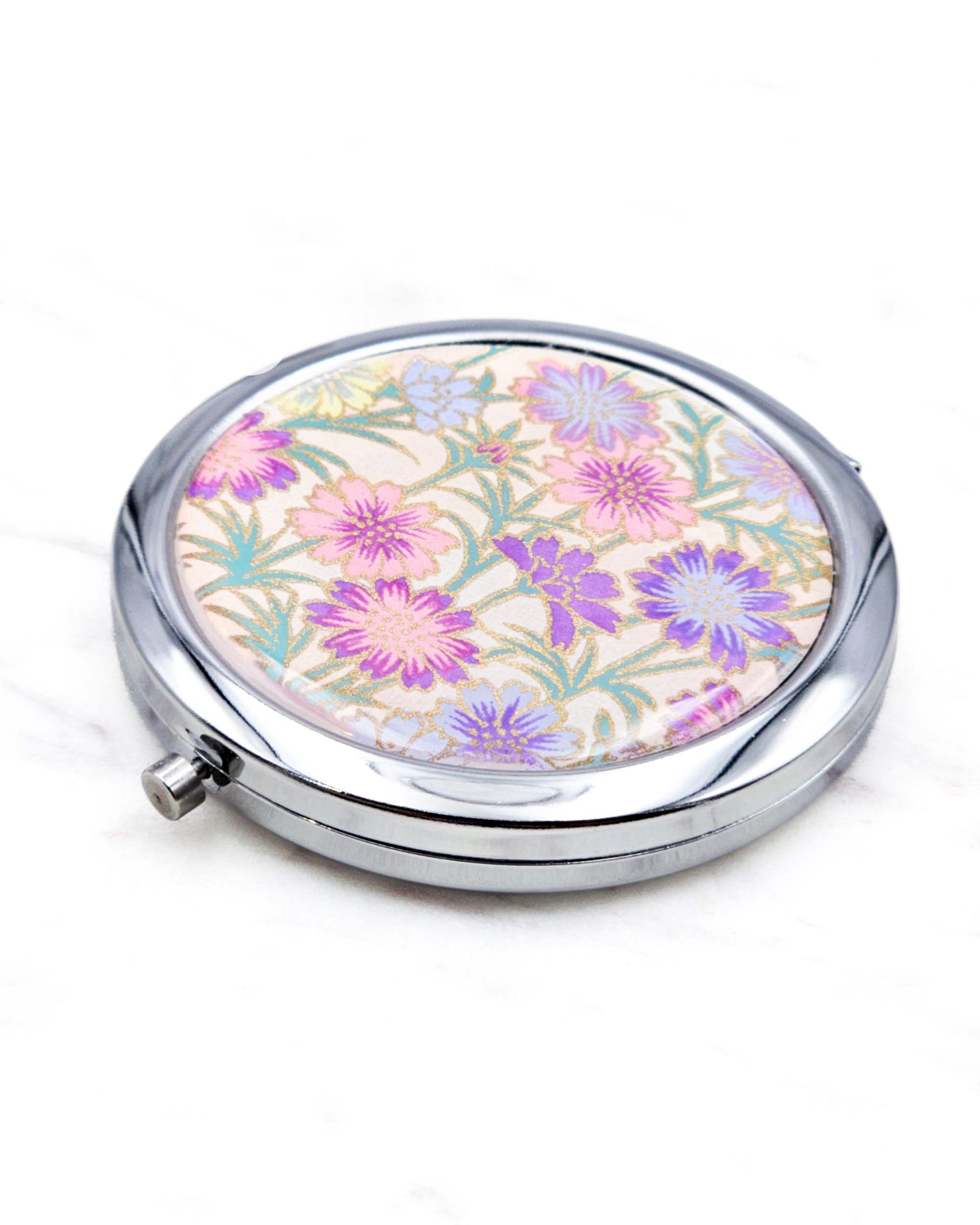 Round silver metal compact mirror featuring purple flower stalks on a light pink background with metallic gold accents.