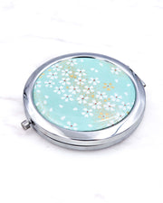 Silver metal compact mirror featuring white cherry blossoms on a turquoise background with metallic gold accents.