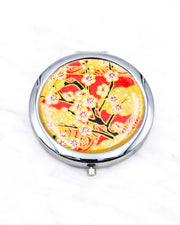 Round silver metal compact mirror featuring white plum blossom branches on a red and gold background with metallic gold accents.