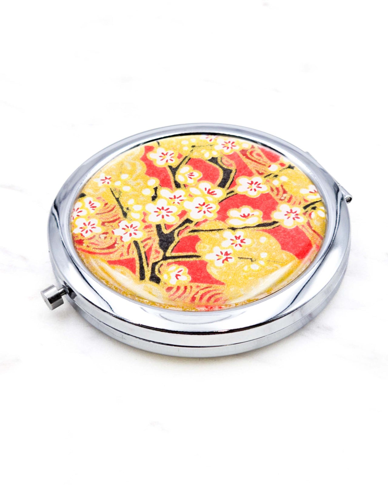 Round silver metal compact mirror featuring white plum blossom branches on a red and gold background with metallic gold accents.