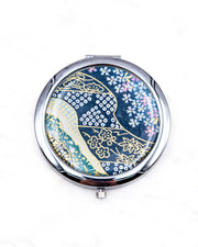 Round silver metal compact mirror featuring pink and white cherry blossoms with assorted traditional Japanese floral and geometric patterns on teal blue background with metallic gold and silver details.