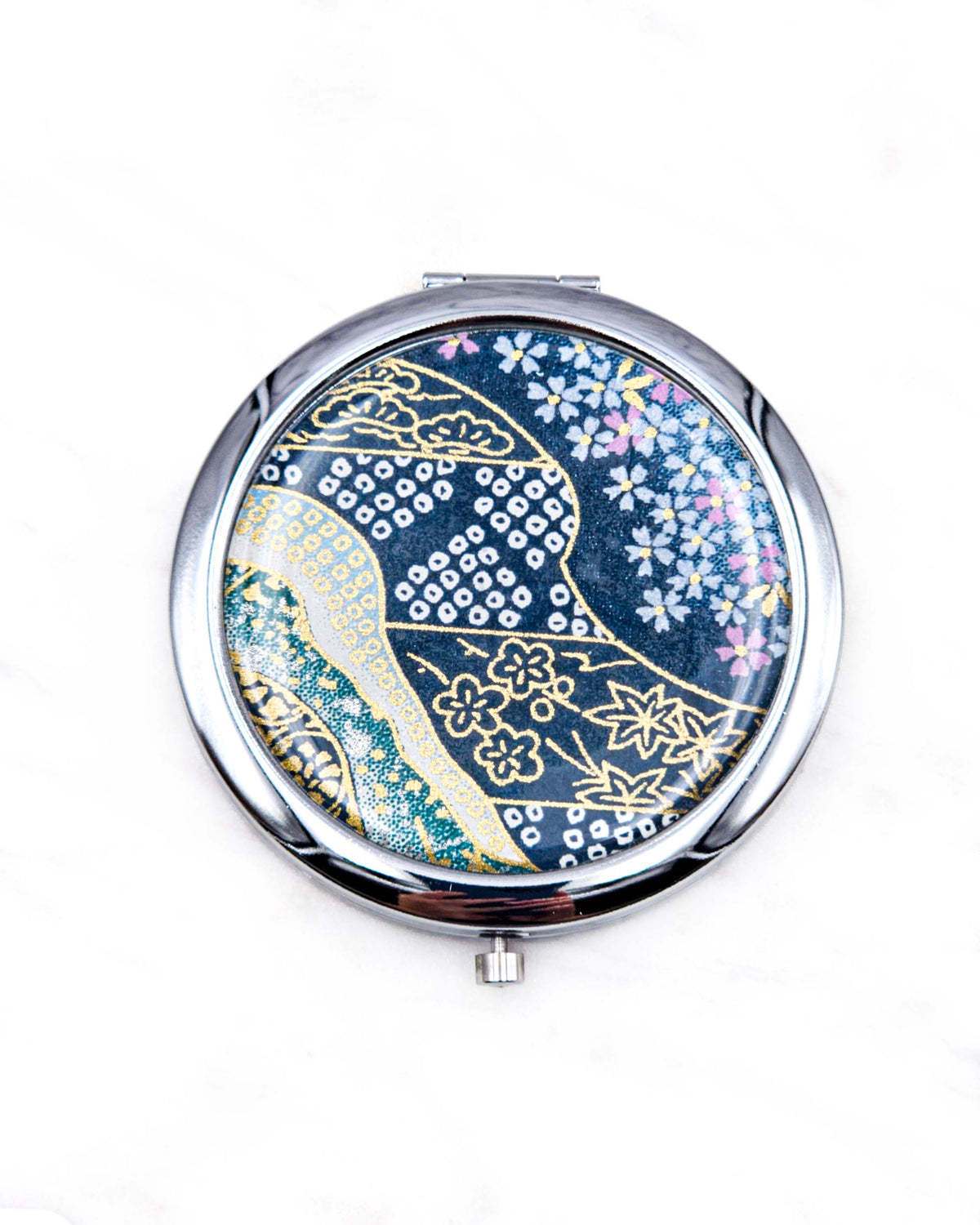 Round silver metal compact mirror featuring pink and white cherry blossoms with assorted traditional Japanese floral and geometric patterns on teal blue background with metallic gold and silver details.