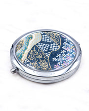 Round silver metal compact mirror featuring pink and white cherry blossoms with assorted traditional Japanese floral and geometric patterns on teal blue background with metallic gold and silver details.