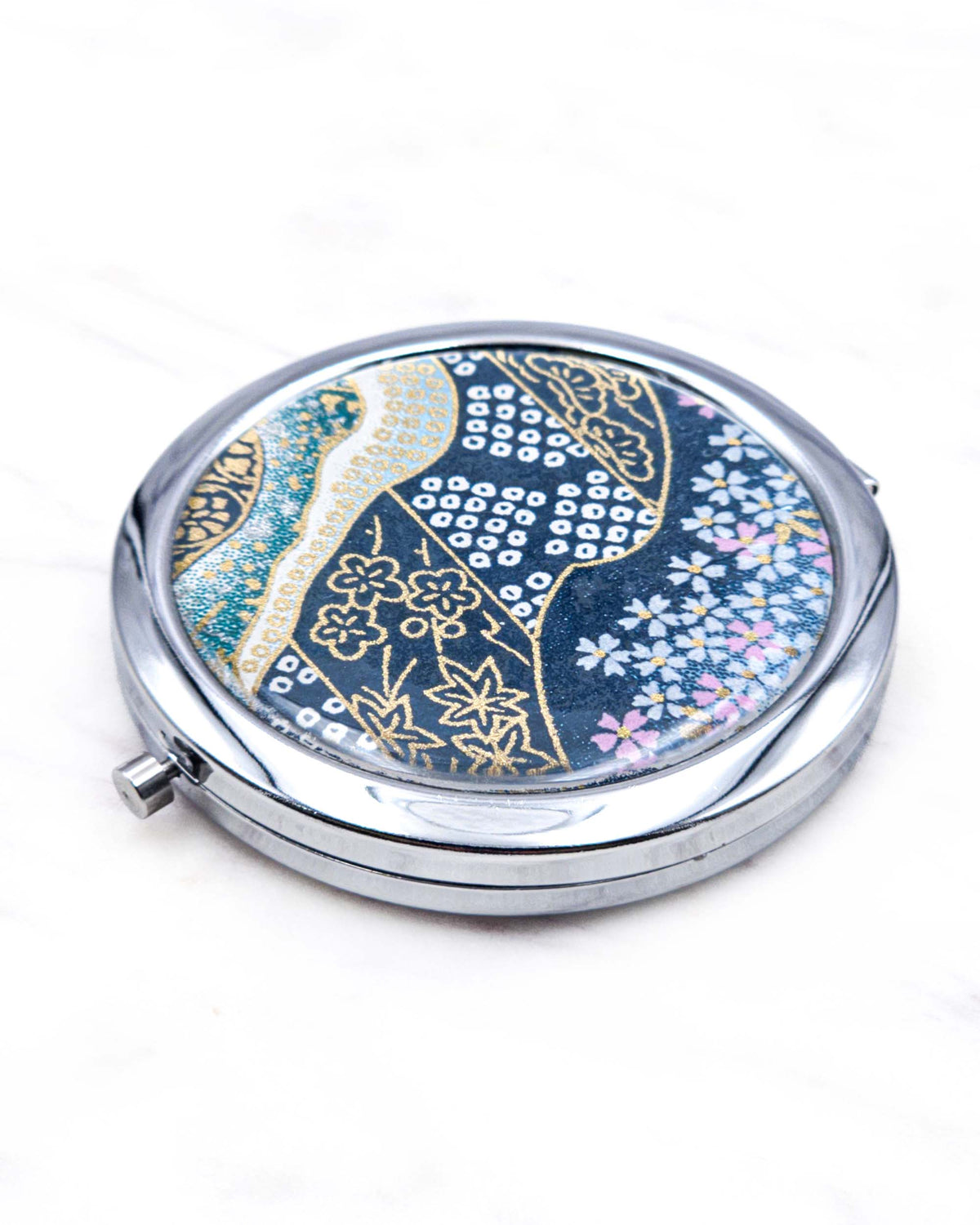 Round silver metal compact mirror featuring pink and white cherry blossoms with assorted traditional Japanese floral and geometric patterns on teal blue background with metallic gold and silver details.