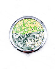 Round silver metal compact mirror featuring white and yellow cherry blossoms on a light green background and a dark green and white wave (aranami) pattern with metallic gold details.