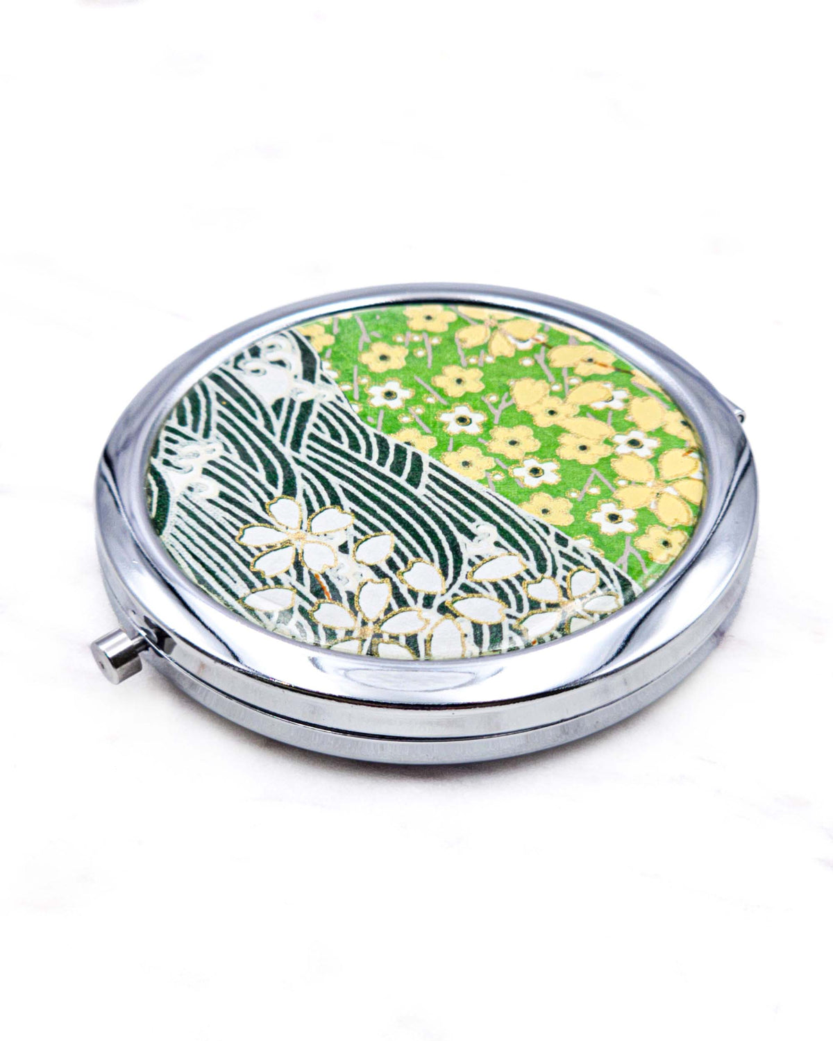Round silver metal compact mirror featuring white and yellow cherry blossoms on a light green background and a dark green and white wave (aranami) pattern with metallic gold details.