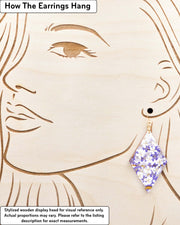 Large wooden diamond shaped earrings featuring purple and white cherry blossoms with metallic gold details, displayed on a stylized wooden head to demonstrate how the earring hangs when worn. Display is not to scale.