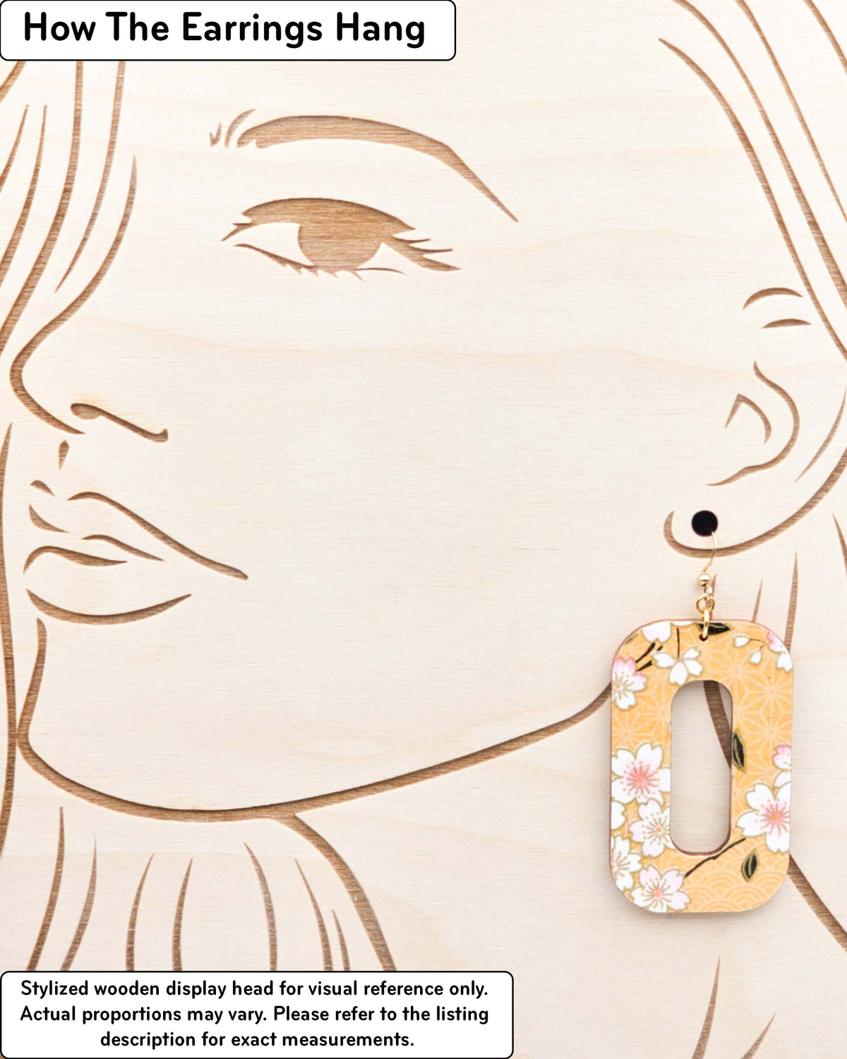 Large wooden hollow rounded rectangular earrings featuring pink cherry blossoms on an orange background with metallic gold details, displayed on a stylized wooden head to demonstrate how the earring hangs when worn. Display is not to scale.