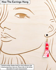 Medium wooden curved bar shaped earrings featuring pink and white pansies with with metallic gold details, displayed on a stylized wooden head to demonstrate how the earring hangs when worn. Display is not to scale.
