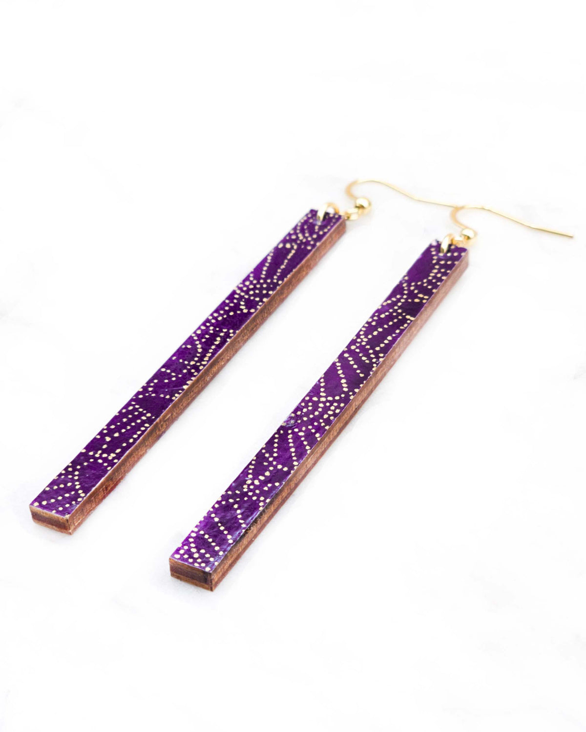 Medium wooden long thin bar shaped earrings featuring small metallic gold pointillistic temari balls on a dark purple background