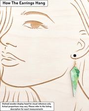 Medium wooden long kite shaped earrings featuring gold, silver, white, and dark green feathery leaves on a green background, displayed on a stylized wooden head to demonstrate how the earring hangs when worn. Display is not to scale.