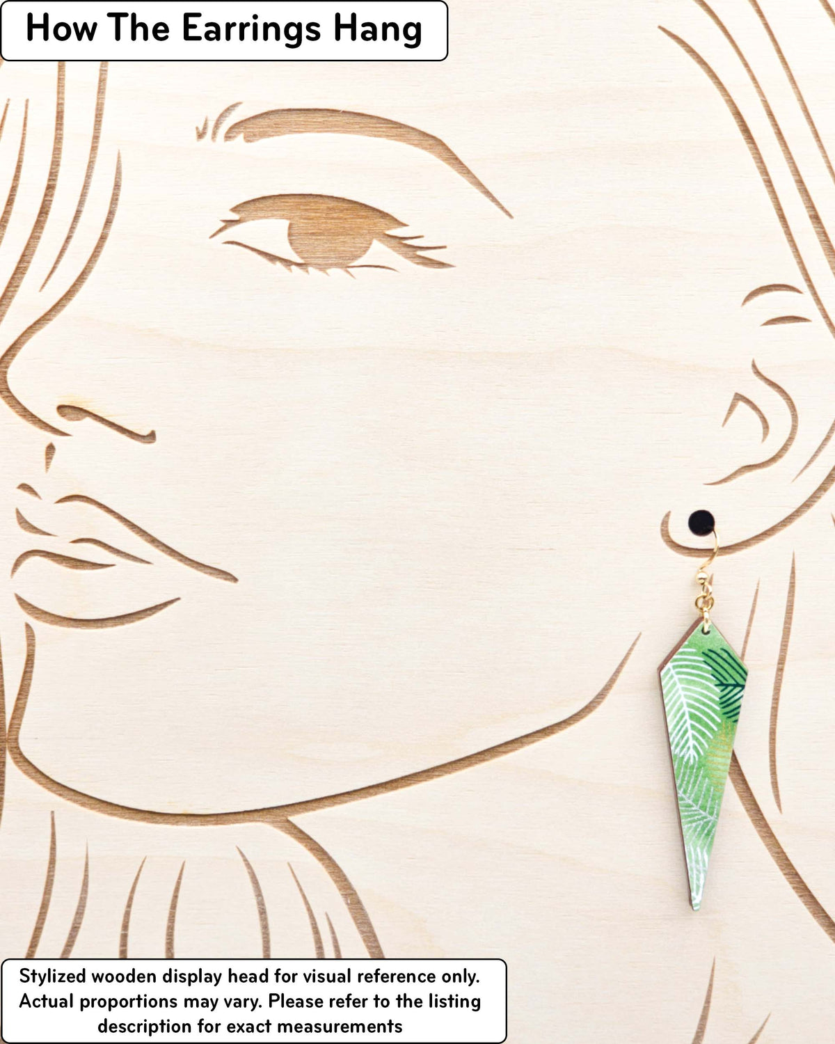 Medium wooden long kite shaped earrings featuring gold, silver, white, and dark green feathery leaves on a green background, displayed on a stylized wooden head to demonstrate how the earring hangs when worn. Display is not to scale.