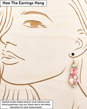 Medium wooden pointed tapered bar shaped earrings featuring pink and beige plum blossoms on a green background with metallic gold details, displayed on a stylized wooden head to demonstrate how the earring hangs when worn. Display is not to scale.