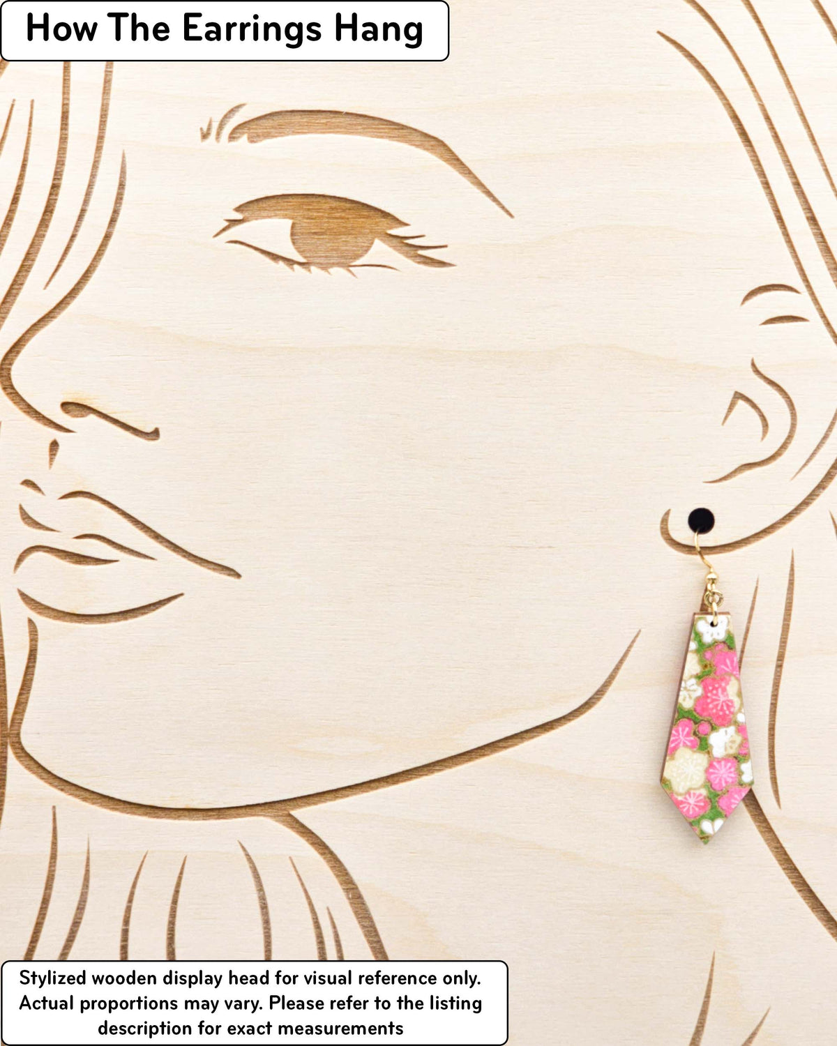 Medium wooden pointed tapered bar shaped earrings featuring pink and beige plum blossoms on a green background with metallic gold details, displayed on a stylized wooden head to demonstrate how the earring hangs when worn. Display is not to scale.