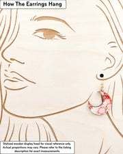 Medium wooden crescent moon shaped earrings featuring red and white cherry blossoms on a red background with metallic gold details, displayed on a stylized wooden head to demonstrate how the earring hangs when worn. Display is not to scale.