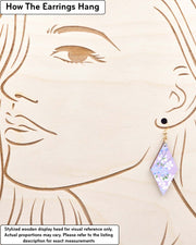 Medium wooden diamond shaped earrings featuring light blue and white cherry blossoms with green leaves and branches on a pearlescent light purple background, displayed on a stylized wooden head to demonstrate how the earring hangs when worn. Display is not to scale.
