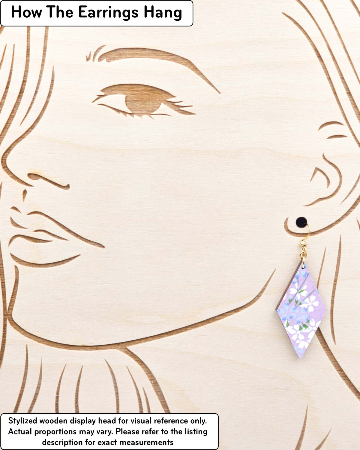 Medium wooden diamond shaped earrings featuring light blue and white cherry blossoms with green leaves and branches on a pearlescent light purple background, displayed on a stylized wooden head to demonstrate how the earring hangs when worn. Display is not to scale.
