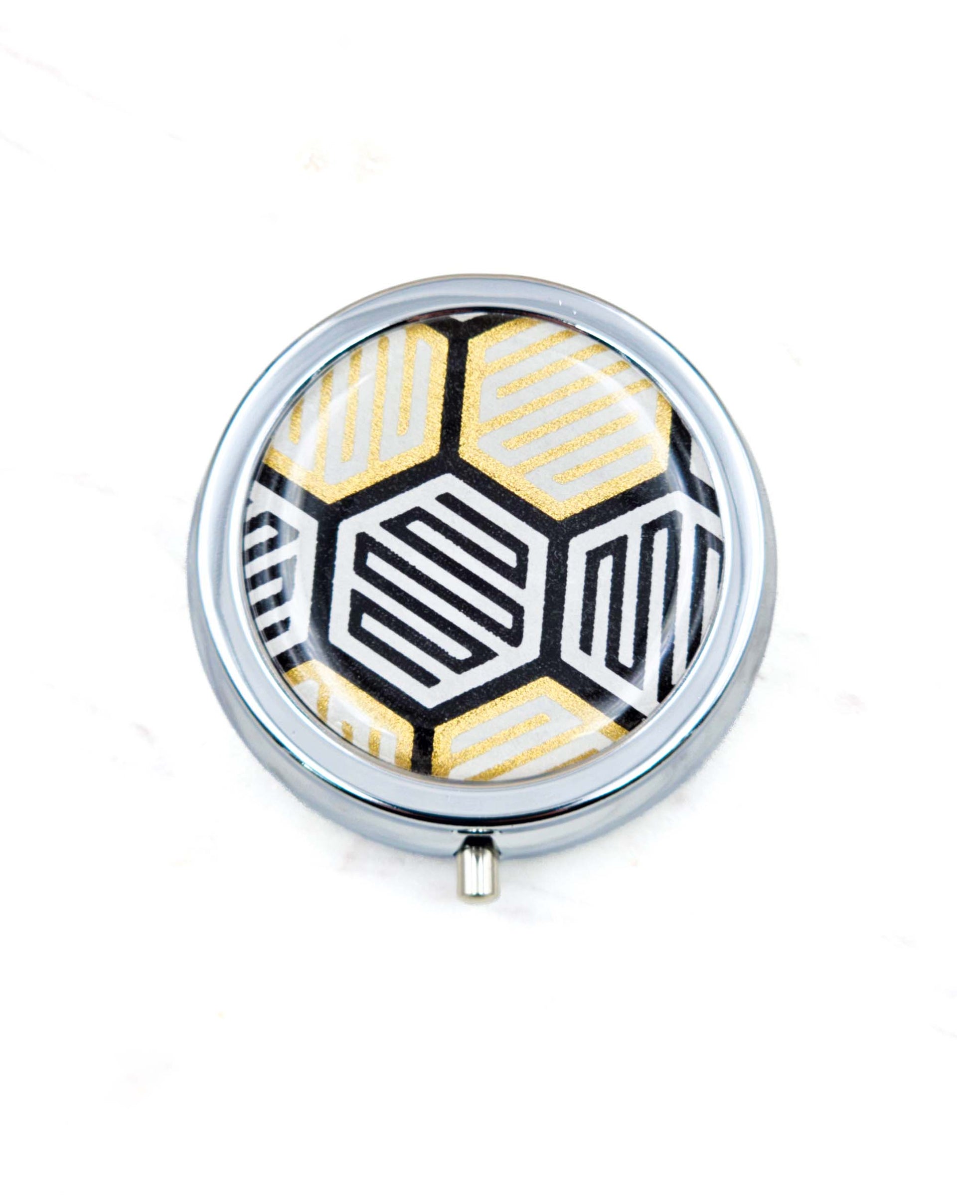 Round silver metal pill box featuring a traditional black, white, and gold kikkou pattern.