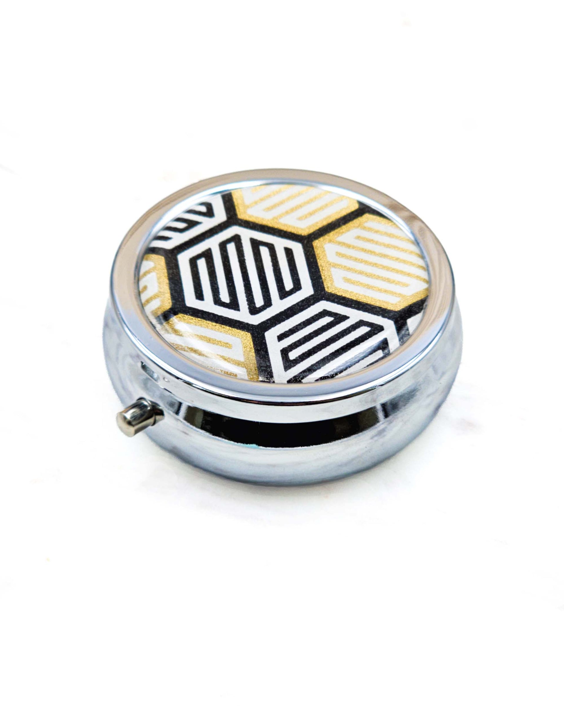 Round silver metal pill box featuring a traditional black, white, and gold kikkou pattern.
