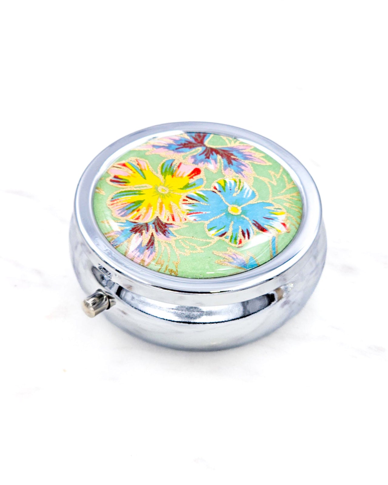 Silver metal pill box featuring blue and yellow flowers on a green background with metallic gold accents.