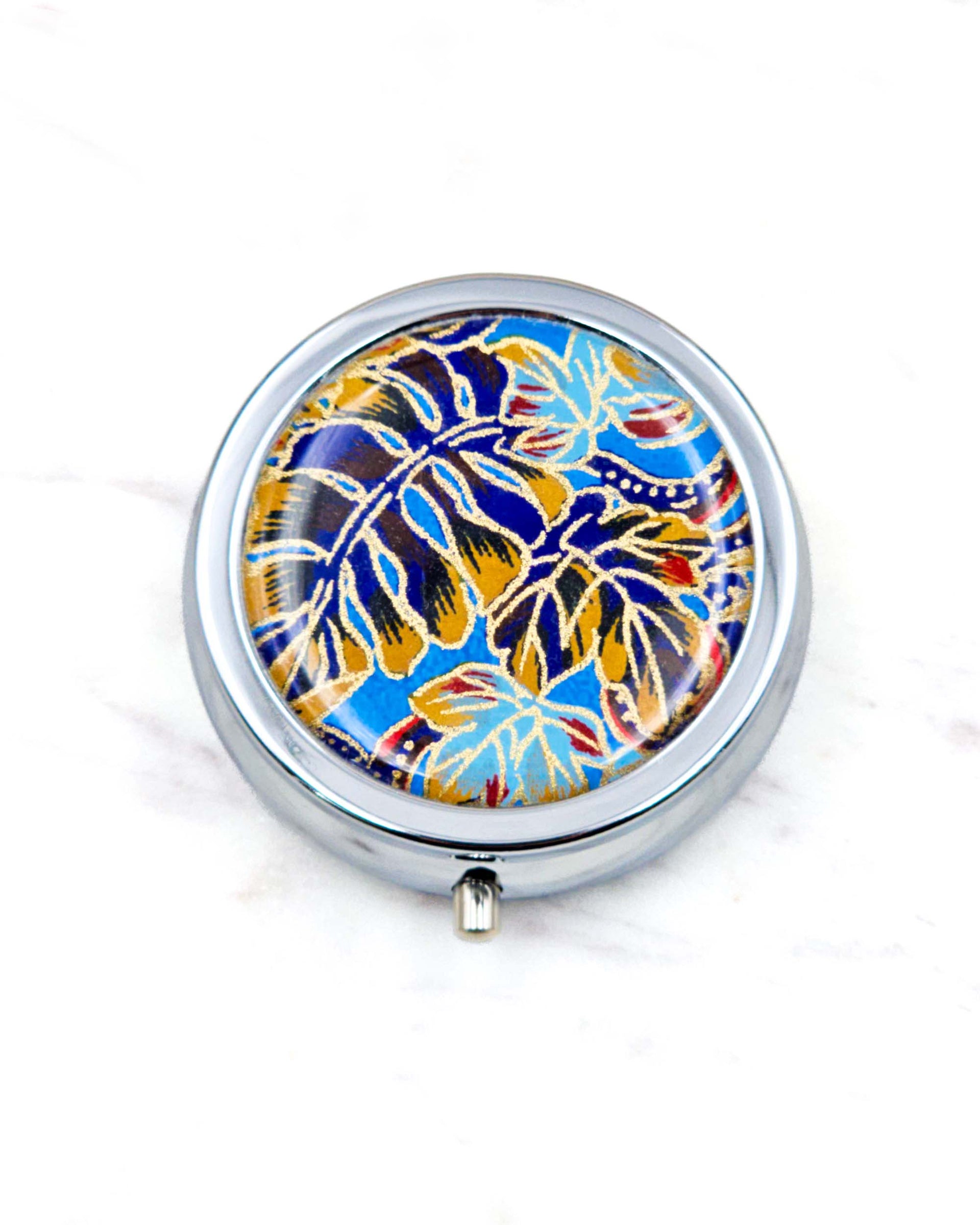 Round silver metal pill box featuring blue, red, and brown frond leaves on a blue background with metallic gold details.
