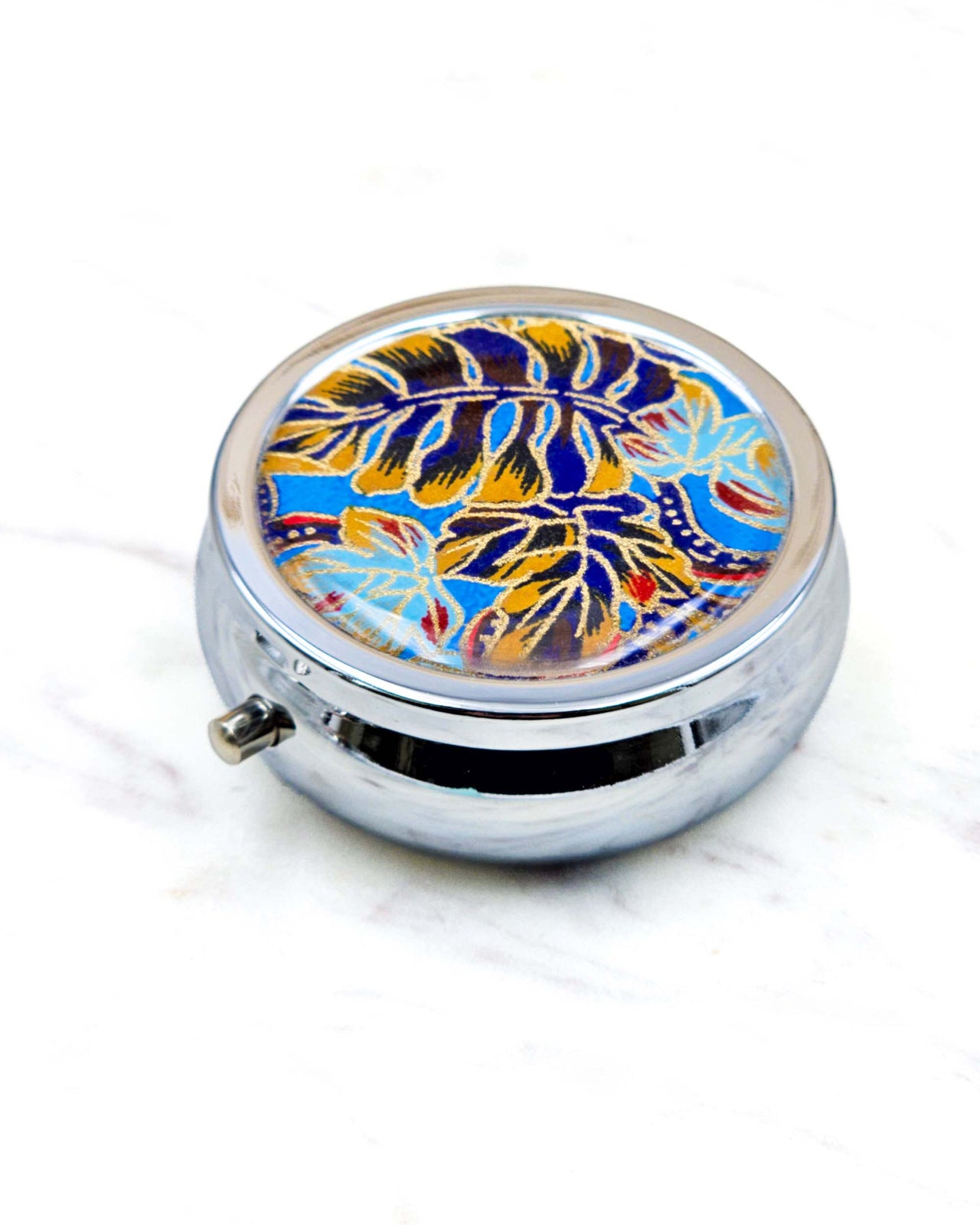Round silver metal pill box featuring blue, red, and brown frond leaves on a blue background with metallic gold details.