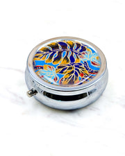 Round silver metal pill box featuring blue, red, and brown frond leaves on a blue background with metallic gold details.