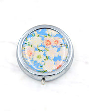 Round silver metal pill box featuring pink and white cherry blossoms on a sky blue background with metallic gold details.