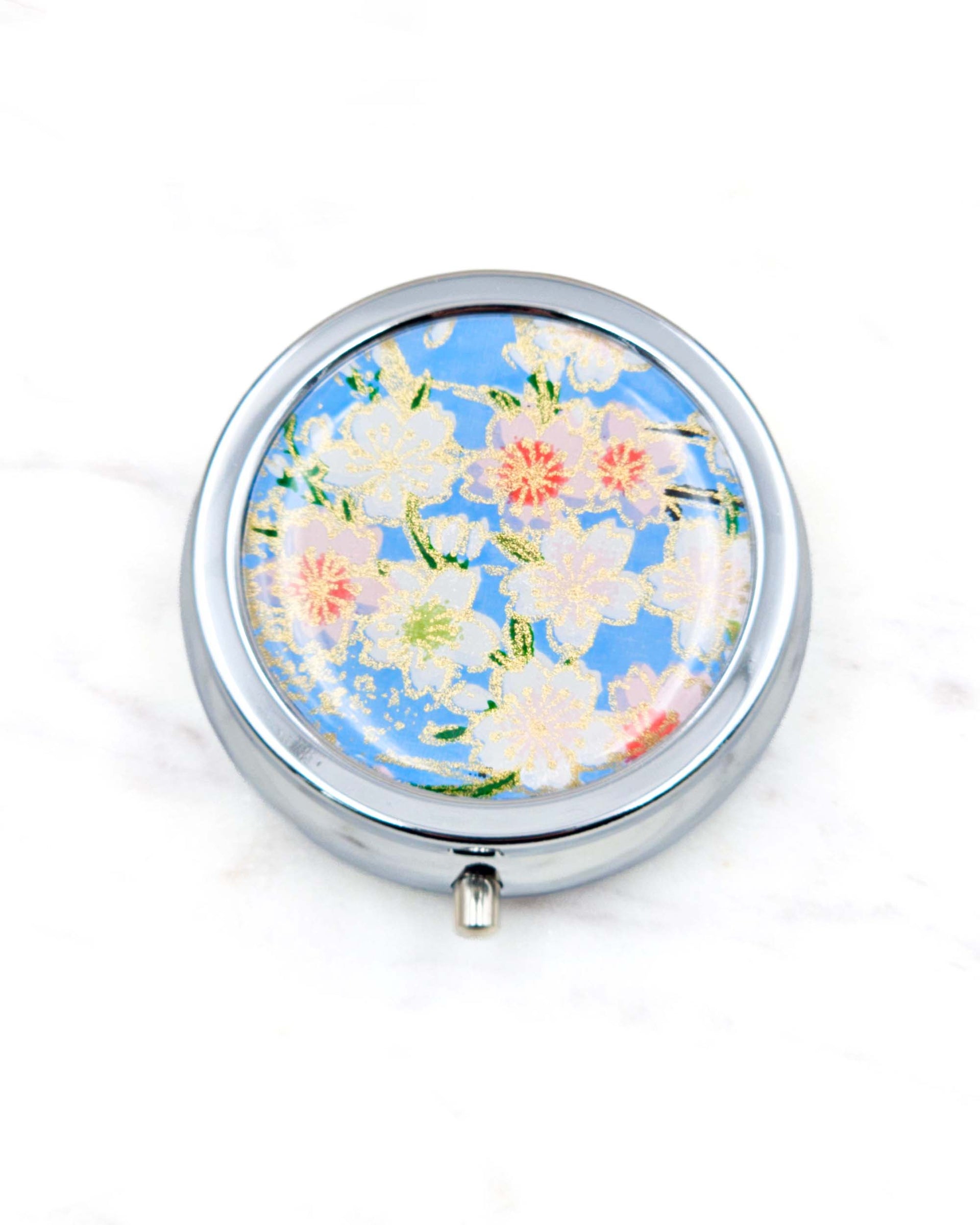 Round silver metal pill box featuring pink and white cherry blossoms on a sky blue background with metallic gold details.