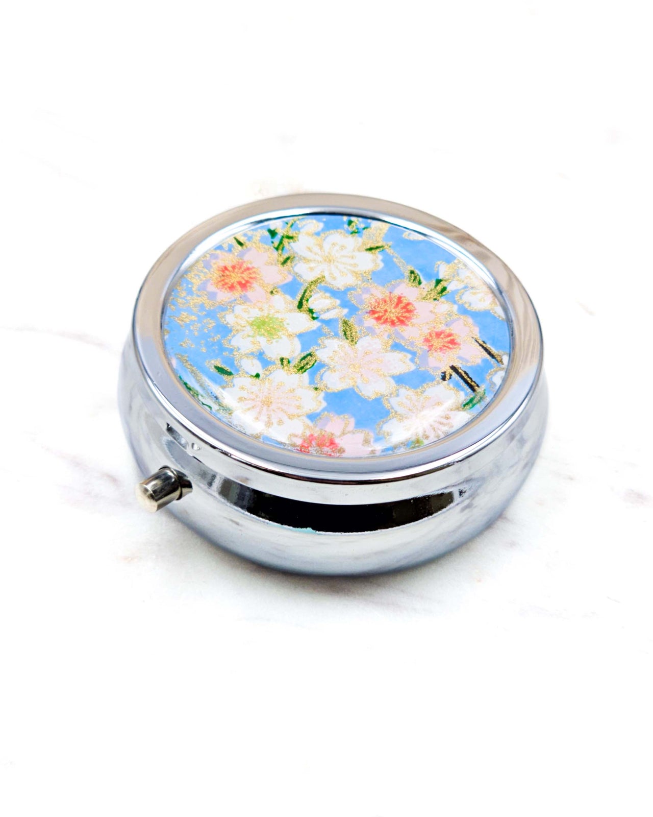 Round silver metal pill box featuring pink and white cherry blossoms on a sky blue background with metallic gold details.